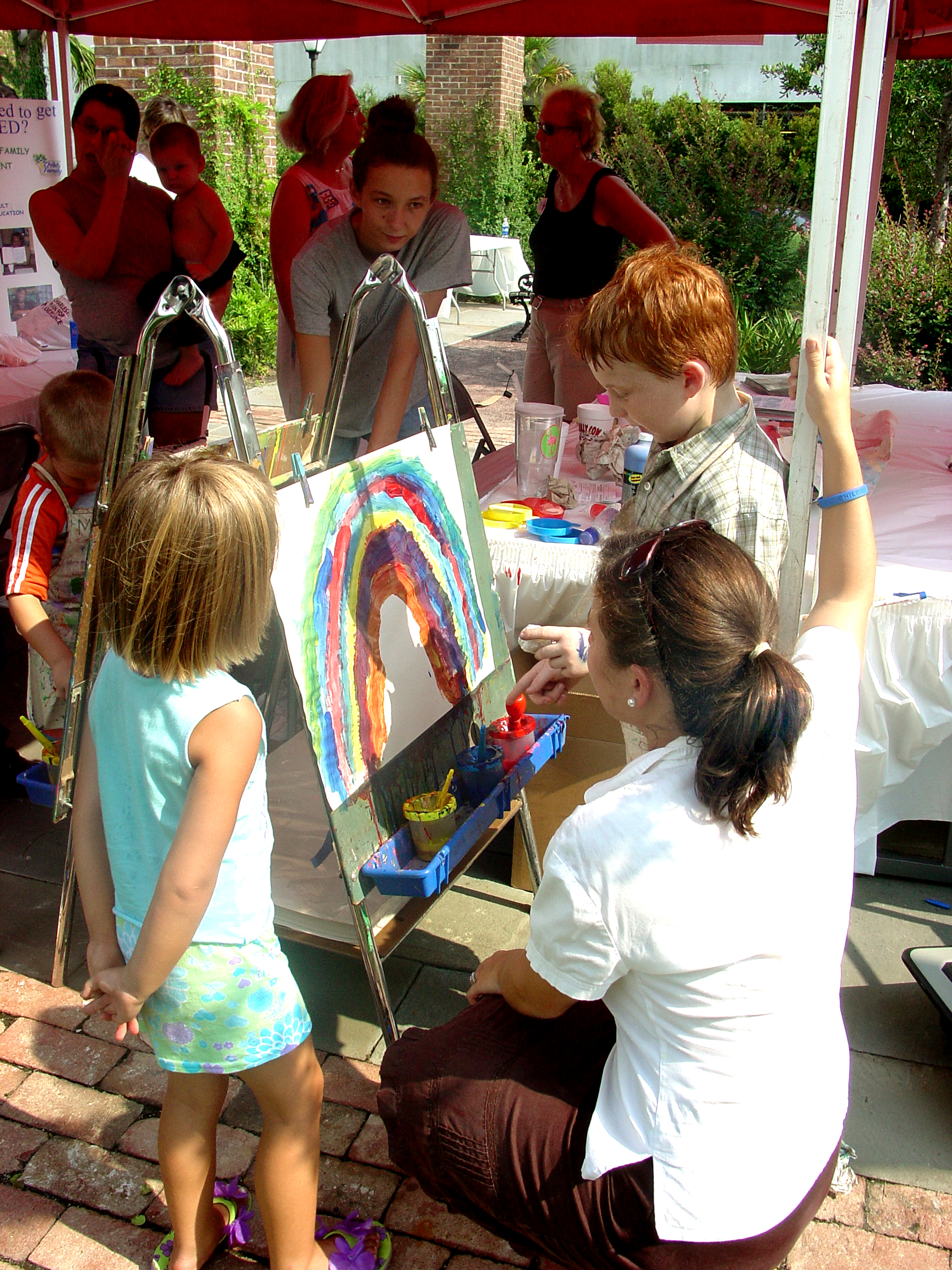 Woman helps children paint a picture of a rainbow at outdoor event